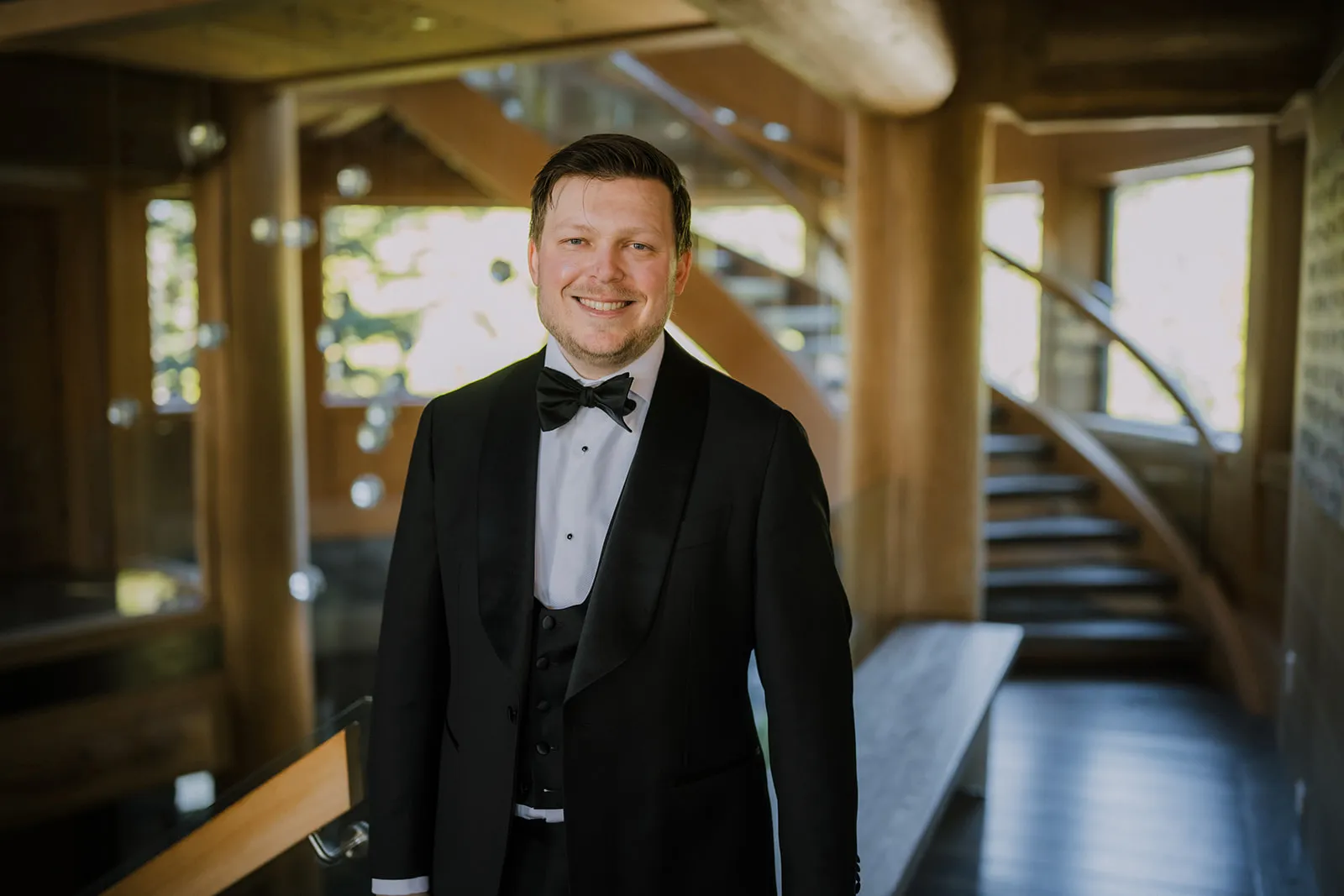 Ready to go James smiling in his black tuxedo and bow tie inside the timber-framed lodge, the sweeping staircase visible behind him