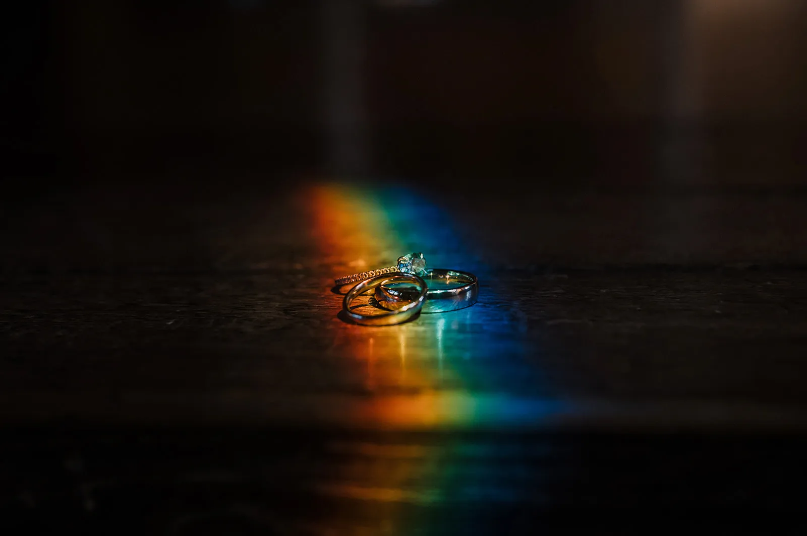 The rings Close-up of the three wedding rings resting on a dark surface, lit by a rainbow of refracted light