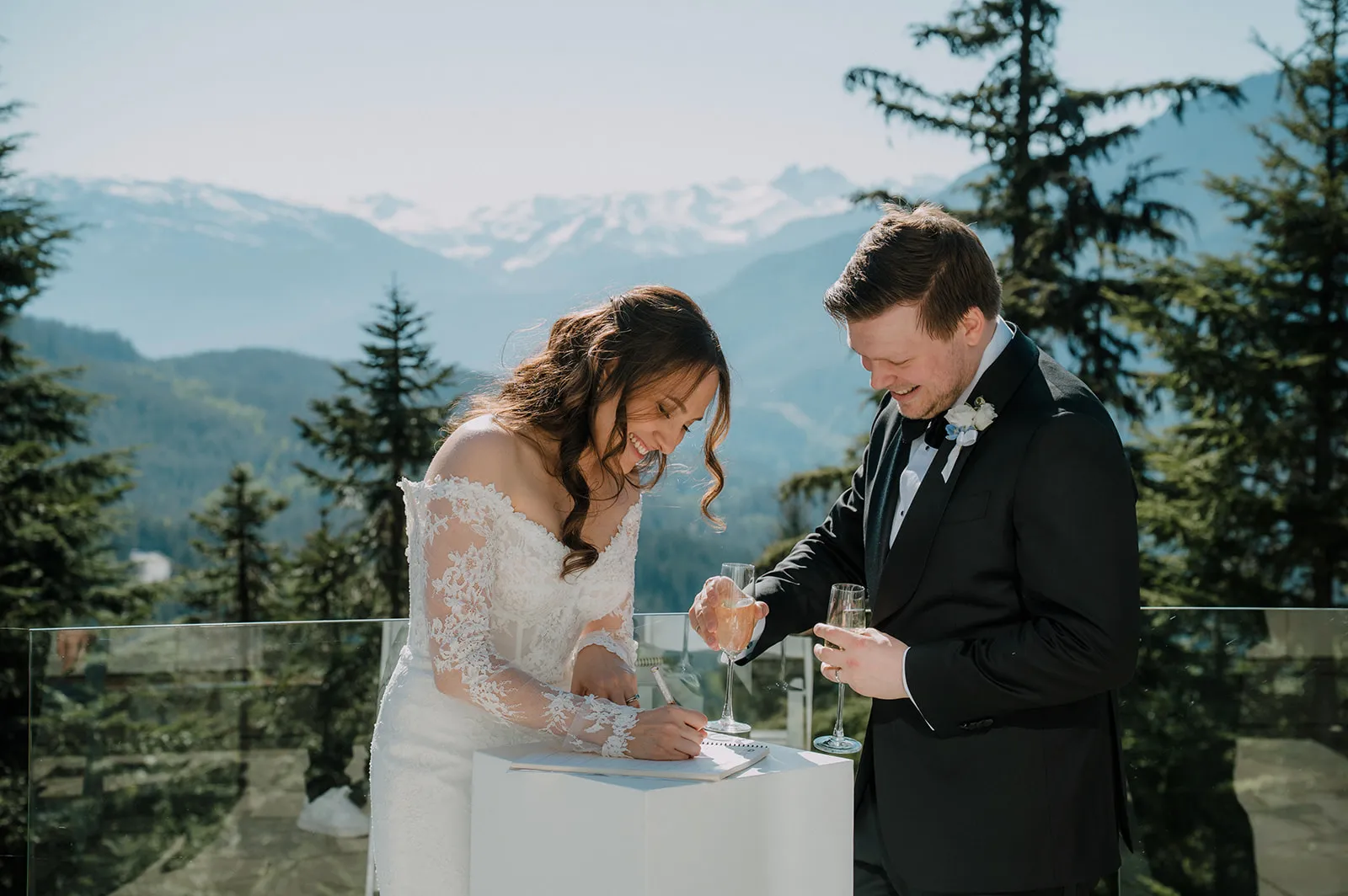 Making it official Both laughing as Bruna signs the register on an outdoor terrace, champagne in hand, with the snow-capped Whistler mountains behind them