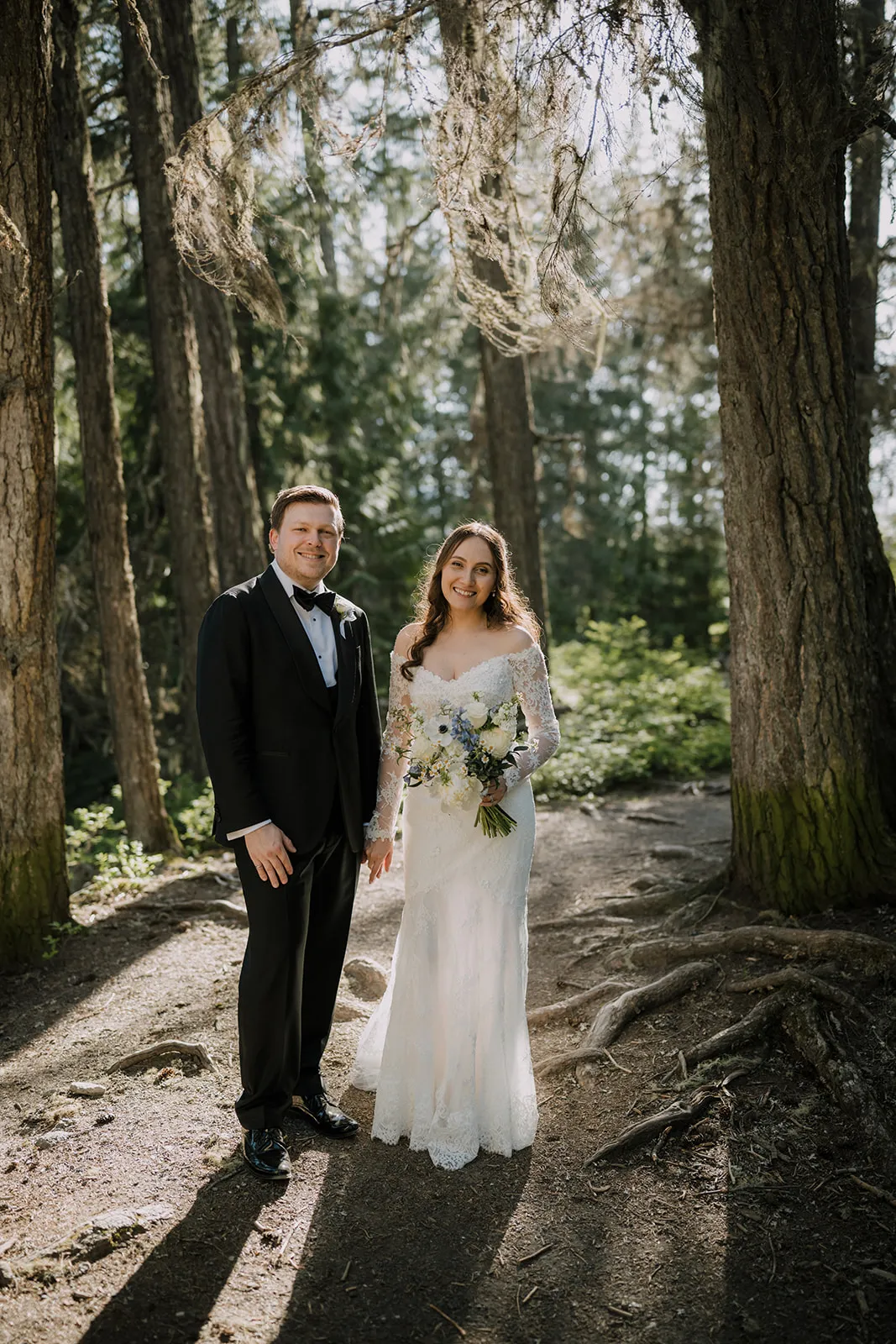Together in the trees James and Bruna standing together on a forest path between tall pine trees, Bruna in a lace off-the-shoulder gown holding a white-and-blue bouquet, James in a black tuxedo